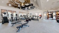 Located in Richardson, Texas, this fitness center offers a spacious, single-floor exercise facility designed to support a comprehensive workout experience. The room is defined by soaring ceilings adorned with a striking sculptural installation of geometric, petal-shaped acoustic panels in black and gold tones, giving the space a modern and visually dynamic character. Recessed lighting and ceiling fans complement the abundant natural light that floods the interior through large, floor-to-ceiling windows lining multiple walls, offering views of the surrounding outdoor environment.

The exercise area is thoughtfully organized to accommodate a wide range of fitness activities. A row of Matrix-brand treadmills and elliptical machines lines the windowed walls, ideal for cardio training with an outdoor view. Cycling bikes, rowing-style machines, and additional cardio equipment are distributed throughout the open floor plan. The strength training section features squat racks with free weights, adjustable benches, a full dumbbell rack, and cable machines, providing options for varied resistance training. A dedicated area along one wall includes stability balls and resistance equipment for functional training. Full-length mirrors on one wall enhance the sense of openness. The light-toned carpeted flooring and crisp white walls contribute to a clean, energizing atmosphere throughout the facility.