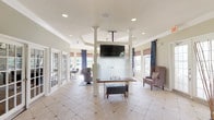 This single-floor community space is designed with an open, welcoming layout that seamlessly connects a central reception or gathering area to distinct lounge and seating zones. The main entry foyer features light-toned tile flooring with diamond inset accents, a pendant lantern fixture, and French glass doors that invite natural light throughout. Crown molding and tray ceilings add architectural refinement, while recessed lighting and stylish orb pendant fixtures provide layered illumination across the space.

At the heart of the interior, white structural columns frame a ceiling-mounted flat-screen television above a frosted glass partition, creating a natural division between the entry corridor and the lounge area. The primary lounge is anchored by a generous sectional sofa adorned with navy and neutral accent pillows, complemented by swivel accent chairs and a glass-topped coffee table. A blue-toned area rug grounds the seating arrangement beneath tray ceilings fitted with additional recessed lighting.

A bank of large windows draped with navy patterned curtains and woven Roman shades lines the rear wall, offering expansive views of an outdoor pool and patio area. A workstation with seating is integrated into this sunlit zone. Throughout, the warm beige color palette, decorative greenery, and carefully chosen furnishings contribute to a polished yet approachable ambiance.