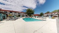 This outdoor amenity space centers around a generously sized swimming pool with clear, blue water, designed for both leisure and lap swimming. The pool is equipped with stainless steel entry rails and is framed by a wide, open concrete sun deck that provides ample room for relaxation and movement around the perimeter.

The surrounding architecture features a distinctive Mediterranean-inspired design, characterized by red clay tile roofing, white stucco walls, and arched entryways. Teal-colored accent pillars are spaced throughout the deck, adding a bold visual contrast to the neutral tones of the hardscape. A covered second-floor balcony with a wood-railed deck overlooks part of the pool area, adding verticality and visual interest to the space.

Lounge chairs and seating arrangements are positioned along both sides of the pool, providing dedicated sunbathing areas. A yellow patio umbrella and additional outdoor furniture offer shaded gathering spots. Mature trees rise above the rooflines, providing natural greenery and partial shade. The space is enclosed by a perimeter fence with gated access points, ensuring security and privacy. Landscape lighting fixtures in the form of classic post lanterns are visible throughout, suggesting the space is also functional and welcoming after dark.