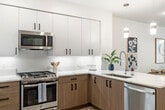 This well-designed one-bedroom, one-bathroom residence offers a thoughtfully laid-out single-floor plan that maximizes functionality and style. The open-concept kitchen flows seamlessly into the living area, featuring two-tone cabinetry with white upper cabinets and warm wood lower cabinets, light quartz countertops, a deep basin sink, stainless steel appliances including a gas range, microwave, and dishwasher, and pendant lighting above the peninsula. Light wood-style flooring extends throughout the main living areas, creating a bright, cohesive aesthetic. The living room is generously proportioned, bathed in natural light from large windows with views of greenery outside, and features clean white walls that complement the modern design. The bedroom offers a comfortable, well-lit retreat with plush carpet, large windows, a dedicated closet, and ample space for furnishings. The full bathroom is finished with subway tile in the tub surround, light tile flooring, a wood-toned vanity, and clean white walls. A private balcony extends the living space outdoors, while a dedicated closet provides additional storage. Clean architectural lines, recessed lighting, and a neutral color palette throughout create a contemporary and inviting atmosphere.