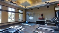 This fitness studio is thoughtfully designed to support a wide range of workout styles within a single, open space. The room features a distinctive coffered ceiling with recessed lighting and a ceiling fan, contributing both visual depth and effective ventilation during intense training sessions. Large windows along one wall allow generous natural light to filter in, creating an inviting and energizing atmosphere while offering views of mature trees outside.

The flooring combines padded mat sections with hardwood-style panels, providing appropriate surfaces for both floor exercises and equipment use. Along the back wall, a dedicated TRX suspension training rig is mounted, accompanied by informational signage and a workout zone display, making it easy for users to follow guided routines. A cubby-style storage unit nearby keeps equipment organized and accessible.

Rowing machines positioned toward the front of the room add a cardiovascular training option, while a stability ball and additional accessories suggest a versatile functional fitness setup. Motivational wall art completes the space, reinforcing an active and focused atmosphere. The overall design balances functionality with a clean, modern aesthetic.