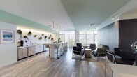 This thoughtfully designed single-floor communal amenity space offers a diverse array of gathering zones, each curated with modern finishes and a cohesive, airy aesthetic. The interior is defined by light wood-toned flooring—transitioning between hardwood-style planks and travertine tile—paired with soft sage-green ceiling accents and crisp white walls that create a calm, welcoming atmosphere throughout.

A well-appointed refreshment bar anchors one side of the space, featuring a sleek white countertop, stainless steel under-counter refrigeration, a coffee station, and a sink. A decorative wall of circular mounted planters with live greenery adds a vibrant, organic touch above the counter. Pendant globe lights hang above a central bar-height island with upholstered stools, ideal for casual gatherings.

A lounge seating area centers around a floor-to-ceiling dark tile fireplace wall, flanked by contemporary low-profile armchairs and a side table. Floor-to-ceiling windows with vertical blinds flood this area with natural light and offer views of the outdoors. Additional seating areas include a tufted sofa beneath a gallery wall of framed artwork, and a compact round dining table with navy upholstered chairs beneath an accent green ceiling.

The space also includes a billiards table and a media wall with a flat-screen display, rounding out the entertainment offerings. Restrooms are conveniently accessible within the space.