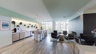 This thoughtfully designed single-floor communal amenity space offers a diverse array of gathering zones, each curated with modern finishes and a cohesive, airy aesthetic. The interior is defined by light wood-toned flooring—transitioning between hardwood-style planks and travertine tile—paired with soft sage-green ceiling accents and crisp white walls that create a calm, welcoming atmosphere throughout.

A well-appointed refreshment bar anchors one side of the space, featuring a sleek white countertop, stainless steel under-counter refrigeration, a coffee station, and a sink. A decorative wall of circular mounted planters with live greenery adds a vibrant, organic touch above the counter. Pendant globe lights hang above a central bar-height island with upholstered stools, ideal for casual gatherings.

A lounge seating area centers around a floor-to-ceiling dark tile fireplace wall, flanked by contemporary low-profile armchairs and a side table. Floor-to-ceiling windows with vertical blinds flood this area with natural light and offer views of the outdoors. Additional seating areas include a tufted sofa beneath a gallery wall of framed artwork, and a compact round dining table with navy upholstered chairs beneath an accent green ceiling.

The space also includes a billiards table and a media wall with a flat-screen display, rounding out the entertainment offerings. Restrooms are conveniently accessible within the space.