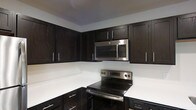 This single-floor space presents a thoughtfully designed interior anchored by a standout kitchen. The kitchen showcases rich, dark espresso-toned cabinetry that extends from floor to ceiling, providing generous storage capacity while delivering a bold, contemporary aesthetic. The deep cabinet tones are elegantly offset by bright white countertops, creating a striking visual contrast that enhances the overall sense of cleanliness and modernity. Stainless steel appliances, including an over-the-range microwave and a smooth-top electric range, complete the kitchen's polished, professional look. The cabinetry features sleek bar-style hardware in a brushed metallic finish, adding a refined detail that ties the space together cohesively. The kitchen is arranged in an efficient layout that maximizes workspace and movement, making it both highly functional and visually appealing. The light-toned walls and ceiling complement the darker elements, keeping the space feeling open and well-balanced. Overall, the space conveys a clean, modern sensibility with attention to detail in its material choices and finishes, making it a practical yet stylish environment suited for everyday living.