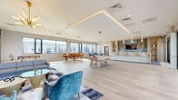 Located at 321 Main St in Hackensack, NJ, this generously sized amenity lounge is a single-floor, open-concept space designed for socializing and relaxation. Light-toned hardwood flooring runs throughout, unifying the distinct activity zones beneath a clean, modern ceiling with recessed LED cove lighting that lends a warm, polished glow.

The lounge area anchors one end of the space, centered around a dramatic floor-to-ceiling marble fireplace wall with a sleek linear gas fireplace and a wall-mounted TV. Plush blue velvet chairs, a contemporary sofa, and a geometric patterned area rug create an inviting gathering spot, while a sculptural gold sputnik chandelier adds a bold design accent.

Adjacent to the lounge, a billiards table serves as a central social focal point, flanked by a dining area with an extended table and modern orange chairs beneath pendant lighting. A stylish wet bar area features a large stone-topped island with white barstools, wood-paneled cabinetry, gold geometric wall accents, and a striking blue agate backsplash.

Floor-to-ceiling glass sliding doors and expansive windows flood the space with natural light and open directly to an outdoor terrace, offering sweeping views of the Hackensack skyline. The terrace features seating and a fire pit, extending the entertaining space outdoors seamlessly.