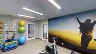 This single-floor fitness space is designed to support active lifestyles in a clean, functional environment. The room is immediately distinguished by a bold, full-wall mural depicting a sweeping horizon landscape in deep blues and warm golden tones, adding a dynamic visual focal point that energizes the space. The walls are finished in a neutral gray, complementing the mural while maintaining a modern, streamlined aesthetic.

Overhead, linear LED strip lighting provides bright, even illumination throughout the room, ensuring excellent visibility for workouts at any time of day. The flooring is a durable, light-toned surface suited for fitness use.

The space is equipped with a stationary cycling bike positioned centrally, ideal for cardio training. Along one wall, a vertical rack holds multiple stability exercise balls in vibrant yellow, green, and blue, adding both functionality and a splash of color to the room.

Two white-framed doors offer access points, one of which features a glass panel revealing a view of a neighboring brick exterior, introducing a hint of natural light. A clearly marked exit sign ensures safety compliance. Overall, the space delivers a practical and visually stimulating fitness environment.