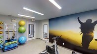 This single-floor fitness space is designed to support active lifestyles in a clean, functional environment. The room is immediately distinguished by a bold, full-wall mural depicting a sweeping horizon landscape in deep blues and warm golden tones, adding a dynamic visual focal point that energizes the space. The walls are finished in a neutral gray, complementing the mural while maintaining a modern, streamlined aesthetic.

Overhead, linear LED strip lighting provides bright, even illumination throughout the room, ensuring excellent visibility for workouts at any time of day. The flooring is a durable, light-toned surface suited for fitness use.

The space is equipped with a stationary cycling bike positioned centrally, ideal for cardio training. Along one wall, a vertical rack holds multiple stability exercise balls in vibrant yellow, green, and blue, adding both functionality and a splash of color to the room.

Two white-framed doors offer access points, one of which features a glass panel revealing a view of a neighboring brick exterior, introducing a hint of natural light. A clearly marked exit sign ensures safety compliance. Overall, the space delivers a practical and visually stimulating fitness environment.