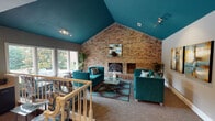 The Estrada Oaks clubhouse is a thoughtfully designed single-story community facility that blends bold architectural character with functional, welcoming spaces. The centerpiece is a striking great room distinguished by soaring vaulted ceilings painted in a rich teal, which contrasts dramatically against the full-height exposed brick wall anchored by a classic wood-burning fireplace. Teal upholstered sofas, a glass coffee table, and layered area rugs create a vibrant yet comfortable seating area, while track lighting and large windows allow natural light to pour in with views of surrounding greenery. The space transitions seamlessly through a split-level layout, with tiled steps leading into the main corridor and common areas. The central hallway serves as a hub, featuring curated artwork, a console display table, and access to multiple zones. A dedicated business nook provides a workstation for residents, while a spacious secondary lounge area is finished with neutral carpet, warm taupe walls, ceiling fan, and tall glass doors that open toward the pool area, filling the room with natural light and outdoor views. Throughout, the clubhouse maintains a cohesive design aesthetic combining earthy neutrals with vibrant teal accents, delivering an inviting and functional gathering environment for its community.