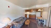 This single-floor unit features an open-concept layout that integrates the living area and kitchen into one cohesive space. The living area is furnished with a patterned sofa and a small oval wood coffee table, positioned atop a dark area rug over hardwood-style flooring that extends throughout the unit. The neutral white walls and textured ceiling keep the space feeling clean and straightforward.

The kitchen runs along the back wall and is equipped with warm-toned oak cabinetry, both upper and lower, providing ample storage. A white electric range, a double-basin sink, and a full-size refrigerator make up the core appliances. Dark laminate countertops contrast with the lighter cabinetry, and a range hood is mounted above the stove. A pendant light fixture hangs near the dining area, where a small table and chairs offer a dedicated eating space.

The overall layout is practical and efficiently organized, making good use of the available floor space. The open design ensures easy flow between the living and kitchen zones, creating a functional environment suited for everyday living.