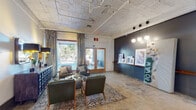 This lobby in Memphis, TN, showcases a compelling blend of industrial heritage and contemporary design. The space is defined by its exposed ceiling with visible ductwork and aged tin detailing, polished and patterned concrete floors, and generous ceiling height that gives the area an open, loft-like feel. Natural light enters through large, curtain-framed glass doors at the front, creating a welcoming entry point. A welcoming lounge area features leather chairs and sofas arranged over a traditional area rug, offering a comfortable gathering spot for residents or visitors. Adjacent to the lounge, a sleek reception desk with a warm wood-topped counter serves as the central point of service. Beside it, a bank of black residential mailboxes is neatly integrated into a deep blue-gray accent wall, adding both functionality and visual structure. Curated artwork, including framed prints and a repurposed street sign, lend the space a locally inspired, artistic character. Globe pendant lights on a track provide focused ambient illumination throughout. A long corridor extends beyond the lobby, leading further into the building, reinforcing the expansive, adaptive-reuse nature of this thoughtfully designed space.