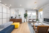 This contemporary single-floor clubhouse is thoughtfully designed to serve as a dynamic community hub. The welcoming entrance lobby sets the tone with a striking tray ceiling, a bold sputnik-style gold chandelier, herringbone-patterned tile flooring, and a central round table that greets visitors warmly. Light wood floors flow throughout, contributing to a bright and cohesive aesthetic.

The lounge area is a standout space, anchored by a floor-to-ceiling tiled fireplace flanked by large windows that invite abundant natural light. Mid-century modern seating, a geometric chandelier, and rich mustard accent window treatments complete the inviting atmosphere.

The dining area opens to an outdoor terrace through glass French doors, offering sweeping views of the surrounding greenery. Geometric-patterned drapes and an angular cage pendant light add a modern design flair.

The kitchen is well-appointed with white upper cabinetry, dark lower cabinets, a white brick-style backsplash, stainless steel appliances, and a spacious island with bar seating — ideal for communal gatherings.

The fitness center features a vibrant geometric mural accent wall in calming blue and green tones, hardwood floors, cardio equipment, and natural light from adjacent spaces.

Accessible, ADA-compliant restrooms with tile wainscoting and grab bars round out the practical amenities of this well-equipped community space.
