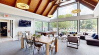 Located at 7999 Wildflower Ln in Columbus, Ohio, this single-floor community clubhouse is a well-appointed gathering space that blends striking architectural design with comfortable, functional interiors. The centerpiece of the space is a dramatic vaulted ceiling with exposed wood beams and tongue-and-groove paneling, which soars overhead in the main living and dining area, creating a warm, lodge-inspired atmosphere. Floor-to-ceiling windows and expansive glass panels line the walls, flooding the interior with abundant natural light and offering sweeping views of the surrounding outdoor area, including a pool and lush greenery.

The open living area features plush sectional seating, a wall-mounted television, and a cozy fireplace set within a clean, white surround — ideal for relaxed gatherings. A large communal dining table with industrial-style metal chairs anchors the dining zone, while a pool table provides additional recreational appeal. Pendant lighting adds warmth throughout.

The kitchen is outfitted with white shaker cabinetry, dark countertops, a classic subway tile backsplash, and stainless steel appliances, including a side-by-side refrigerator and dishwasher. A breakfast bar with seating connects the kitchen to the main space. Light hardwood flooring runs through the entrance and kitchen, transitioning to plush carpet in the main gathering areas. Restroom facilities and a hallway complete this inviting, versatile space.