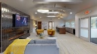 Located at 249 Crittenden Way in Rochester, NY, this thoughtfully designed single-floor space offers an open, flowing layout that combines comfort and modern elegance. Soaring vaulted ceilings with exposed decorative beams define the architectural character throughout, lending an airy, expansive feel to every area.

The living lounge area serves as a central gathering point, anchored by a striking floor-to-ceiling fireplace clad in dark glossy tile and topped with a mounted flat-screen display. Bold gold velvet accent chairs and a plush gray sectional sofa surround a sculptural glass coffee table, all set atop patterned carpeting with warm yellow accents. A statement cloud-form pendant light illuminates the space above.

Adjacent to the lounge, a well-appointed kitchenette and bar area features a wood-toned island with bar seating, integrated cabinetry, and beverage refrigeration — ideal for hosting and entertaining.

The dining area is bathed in natural light from multiple large windows offering outdoor views, and is crowned by a stunning capiz shell chandelier. A round white marble-top table paired with rich burgundy velvet chairs creates an inviting setting for gatherings.

Throughout the space, recessed lighting, neutral wall tones, and cohesive design elements tie each zone together into a polished, welcoming environment.