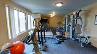 This fitness room offers a practical and functional workout space designed to accommodate a variety of exercise routines. The room features durable rubberized flooring, providing a safe and supportive surface for strength and cardio training alike. Overhead flush-mount light fixtures illuminate the space evenly, while a row of windows along one wall allows natural light to filter in, creating a more open and welcoming atmosphere.

The room is equipped with a solid selection of cardio machines, including an elliptical trainer, a stationary bike, and a treadmill positioned toward the back. A dedicated free weights area includes a tiered dumbbell rack stocked with a range of colorful weights, accompanied by an adjustable workout bench for strength exercises. A multi-station cable machine is positioned along the far wall, offering a wide range of resistance training options.

A wall-mounted air conditioning unit helps maintain a comfortable temperature during workouts. A large mirror on the far wall visually expands the space and allows users to monitor their form. The overall layout is efficient, making good use of the available space while keeping equipment accessible and organized.