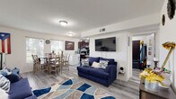 Located at 1207 Palm Glades Dr in Belle Glade, Florida, this single-story home offers a practical and well-organized layout designed for everyday living. The open-concept main area seamlessly connects the living room, dining space, and kitchen, creating a cohesive and functional environment throughout.

The living area is anchored by light wood-style laminate flooring that extends throughout the home, providing a clean and consistent aesthetic. A bold blue sofa set paired with a patterned area rug in blue and neutral tones adds color and warmth to the space. Natural light enters through front-facing windows, brightening the room alongside flush-mount ceiling lighting.

The dining area accommodates a full-sized wooden table with seating for multiple guests, positioned adjacent to the kitchen for convenient flow. The kitchen features rich dark mahogany cabinetry, granite-style countertops, black appliances including a range and refrigerator, and a window above the sink that brings in additional natural light.

The bathroom is neatly appointed with a white vanity, a tub-shower combination, and wood-style flooring consistent with the rest of the home. Overall, the space presents a straightforward, livable layout suited for comfortable residential use in South Florida's Belle Glade community.