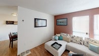 This single-floor residence is thoughtfully organized across several distinct living areas, each with its own character and function. The living room serves as the heart of the home, featuring warm wood-look flooring and a notable terracotta-toned accent wall that adds depth and warmth to the space. Natural light filters in through blinds on multiple windows, enhancing the welcoming atmosphere. A sectional sofa and accent chair provide comfortable seating, while wall art and decorative touches contribute to the cohesive design.

Adjacent to the living area, the dining space accommodates a wooden dining table with matching chairs, positioned near vertical blinds that allow for adjustable natural light. A decorative wall-mounted wine rack adds a personalized detail to this area.

The kitchen features speckled countertops, dark upper cabinetry, and essential appliances, including a refrigerator. A tall utility panel provides additional storage and functionality within the space.

The bedroom is carpeted and generously sized, with ample room for a full bedroom suite including an ornate bed frame, a large dresser with mirror, and a nightstand. Windows invite natural light, contributing to a bright and restful environment.

The bathroom includes a modern gray vanity with an integrated sink, a wall-mounted mirror with lighting above, and standard fixtures, presenting a clean and practical layout.