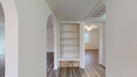 This single-floor residence offers a well-organized layout with consistent wood-look flooring running throughout all living areas. The entryway welcomes you with a clean, open hallway equipped with a thermostat and direct access to the main living spaces. A standout architectural detail is the elegant arched doorway that connects areas of the home, complemented by a built-in wall bookshelf featuring a decorative stone base — adding character to the interior. The kitchen is designed with a pass-through breakfast bar that opens to the main living and dining area, creating a connected and sociable layout. White cabinetry, ample counter space, and black appliances including an electric range and overhead exhaust hood provide a functional cooking environment. The living area benefits from natural light through a large sliding glass door fitted with vertical blinds, offering outdoor views. Bedrooms are well-proportioned, each featuring ceiling fans with light fixtures and windows with blinds. One bedroom includes a walk-in closet for added storage convenience. The bathroom is fitted with a tub-shower combination surrounded by clean, large-format white subway tile and chrome fixtures. Textured ceilings are found throughout, adding a subtle design element to this move-in ready home.