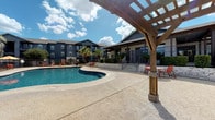 Republic Woodlake, located in San Antonio, TX 78249, presents a thoughtfully designed community clubhouse and leasing office that blends industrial aesthetics with eclectic, upscale furnishings. The interior showcases soaring exposed concrete ceilings with visible ductwork and track lighting, dark hardwood floors, and expansive floor-to-ceiling windows that flood the space with natural light and offer open views of the surrounding landscape. The welcoming lobby features a curved reception desk, patterned accent walls, and a mix of distinctive seating including wicker egg chairs and ornate lounge furniture. A spacious lounge area is appointed with richly layered Persian-style rugs, vintage-inspired armchairs, leather ottomans, and sheer curtain panels that frame the panoramic glazing. The interiors flow seamlessly toward additional gathering areas highlighted by warm pendant lighting. Outside, the resort-style pool serves as a central amenity, featuring a curvilinear design surrounded by a generous sundeck with lounge seating. A substantial wood pergola anchors the poolside area, providing shaded gathering space. The multi-story residential buildings, finished in deep navy and charcoal tones, frame the courtyard and are accented by mature palm trees and lush landscaping, creating a vibrant outdoor retreat under the expansive Texas sky.