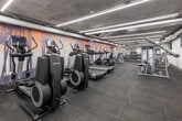 Located at 50 Portland Street in Toronto's vibrant King West neighbourhood, this fitness facility offers a well-appointed, single-level space designed to support a comprehensive workout experience. The main gym area is generously proportioned, featuring a row of Life Fitness cardio machines — including treadmills, ellipticals, and stationary bikes — arranged along one wall, leaving ample open floor space for movement. A full selection of strength equipment, including cable machines, resistance stations, and a dumbbell rack, is distributed throughout the room. The exposed concrete ceiling lends an industrial character to the space, while linear LED strip lighting provides bright, even illumination across the rubber-tile flooring. A striking nature-inspired mural — depicting tall trees against a warm, sunset-toned sky — runs the length of one wall, adding visual interest and a sense of calm to the energetic environment. A separate studio area is accessible from the main floor, featuring herringbone wood-look flooring, full-length mirrors, ballet barres, and a bold geometric accent wall in orange and white. This dedicated studio space is well-suited for yoga, stretching, barre, or group fitness classes, rounding out a versatile and thoughtfully designed amenity facility.