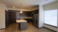 This single-floor space showcases a thoughtfully designed open-concept kitchen that serves as the focal point of the layout. The kitchen features dark espresso-toned cabinetry that extends to the upper walls, providing generous storage capacity. A colorful mosaic tile backsplash adds a decorative accent between the upper and lower cabinets, introducing a subtle pop of visual interest against the darker tones. The countertops are finished in a clean, light-colored surface that contrasts elegantly with the cabinetry.

A central kitchen island with a matching dark base and light countertop offers additional workspace and casual dining potential. Stainless steel appliances, including a French door refrigerator, gas range, and microwave, lend a polished, contemporary feel. Overhead, a combination of pendant lighting above the island and a track lighting system ensures the kitchen is well-lit and functional.

The warm hardwood-style flooring flows throughout, contributing to a cohesive and inviting atmosphere. Natural light enters through a window fitted with white shutters, adding brightness to the space. The overall design balances modern aesthetics with practical functionality.