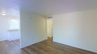 This single-floor space presents a clean and minimalist aesthetic, characterized by crisp white walls and warm-toned hardwood flooring that runs throughout. The interior maintains a bright, airy feel, enhanced by the light-colored walls and smooth white ceilings. A centrally placed doorway connects the main room to an adjacent area, providing a sense of depth and connectivity within the layout. The ceiling-mounted HVAC vent indicates a modern climate control system integrated seamlessly into the space. The rooms are well-proportioned, offering a comfortable sense of openness without feeling overwhelming. The overall ambiance is neutral and inviting, making it a versatile space suited to a variety of uses. The consistent flooring and color palette throughout create a cohesive and unified interior environment. Simple electrical outlets are positioned thoughtfully along the baseboards, maintaining the clean lines of the walls. The white-paneled interior door visible through the doorway adds a classic touch to the otherwise understated design. The space is move-in ready, with a timeless, no-fuss interior that provides a strong foundation for personalization.