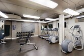Located at 510 21st St NW in Washington, DC, this fitness center offers a well-equipped exercise space designed to support a comprehensive workout routine. The room features durable rubber flooring throughout, providing a safe and practical surface for a variety of exercises. Overhead, suspended fluorescent light panels ensure the space is brightly and evenly illuminated.

The cardio section is outfitted with multiple treadmills, elliptical machines, and stationary and spin bikes — several of which are equipped with personal display screens for entertainment or tracking. A strength-training area includes a multi-function cable machine, an adjustable weight bench, and a full rack of dumbbells in varying weights. Medicine balls and a flat bench round out the free-weight offerings. Wall-mounted mirrors expand the visual sense of space and allow users to monitor their form during workouts. A mounted flat-screen television adds an entertainment option for those on cardio equipment. A water cooler is conveniently positioned within the room. Motivational athletic artwork adorns one wall, adding character to the otherwise clean, utilitarian aesthetic. Exposed infrastructure at the ceiling gives the space an industrial edge, consistent with its functional purpose.