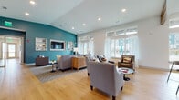 This single-story community clubhouse is thoughtfully designed to offer residents a range of amenity spaces within a cohesive, contemporary aesthetic. Warm-toned hardwood flooring flows throughout, connecting each area seamlessly while vaulted ceilings lend an open, airy quality to the interior.

The central lounge serves as the heart of the space, anchored by a striking teal accent wall that houses a wall-mounted flat-screen television flanked by built-in display shelving. Comfortable gray upholstered seating is arranged around a central coffee table on a textured area rug, while large windows dressed in sheer curtains invite abundant natural light and views of the surrounding community courtyard.

A dedicated coffee and kitchenette bar features sleek navy cabinetry, a light quartz countertop, a self-serve beverage station, and a full-size refrigerator, all set against an eye-catching textured tile accent wall. Abstract framed artwork punctuates the corridors and transition spaces, adding a gallery-like character throughout.

The mailroom provides organized package and mail access with banks of black mailboxes beneath pendant lighting. The fitness center is well-equipped with strength and cardio equipment, floor-to-ceiling mirrors, rubber flooring, and large windows that maintain a bright, motivating environment accented by a teal tray ceiling.