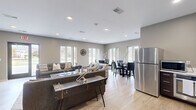 Located at 1633 W Bender Rd in Glendale, Wisconsin, this single-floor communal space offers a thoughtfully designed layout combining a living room and dining area in a cohesive, open environment. The interior is finished with light hardwood flooring throughout, neutral gray and taupe wall tones, and recessed lighting that creates a clean, contemporary aesthetic.

The living room is a welcoming gathering space anchored by a wall-mounted flat-screen television set against a bold accent wall. Comfortable seating arrangements, including a plush sofa and accent chairs, are paired with a substantial coffee table and area rug to define the space. Large windows along the perimeter flood the room with natural light and provide pleasant views of the surrounding greenery.

The dining area features a round glass-top table with sleek black leather chairs, positioned near a generous bank of windows that frame outdoor views and enhance the sense of openness. A stainless steel refrigerator is conveniently accessible nearby, adding functional utility to the space.

Throughout, the design strikes a balance between modern style and comfortable functionality, with consistent finishes, recessed lighting, and an abundance of windows that connect the interior to the outdoors. The overall ambiance is bright, neutral, and inviting — well-suited for communal gathering and everyday use.