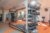 Located at 1770 Peachtree St NE in Atlanta's Midtown neighborhood, this fitness center offers a bright and modern workout environment designed to energize and inspire. Floor-to-ceiling glass curtain walls line the perimeter, flooding the space with abundant natural light and framing panoramic views of the surrounding Atlanta skyline and lush tree canopy below — a distinctive backdrop that sets this facility apart.

The open floor plan is anchored by a central cylindrical column and features vibrant orange-and-red patterned carpet flooring that adds energy to the space. The cardio zone is generously equipped with a row of Life Fitness treadmills, elliptical cross-trainers, and stationary bikes, all positioned to take advantage of the sweeping outdoor views. Overhead, circular pendant light fixtures provide warm, even illumination throughout, complemented by large industrial fans mounted on the walls to maintain airflow.

On the opposite end of the floor, a dedicated strength-training area offers a multi-station cable machine, a full rack of dumbbells, resistance equipment, and a functional training turf lane with agility markings. Mirrors line portions of the walls to support proper form and enhance the sense of space. The overall ambiance is clean, modern, and motivating — a professional-grade fitness environment suited for a comprehensive workout routine.