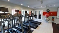 This single-floor fitness center offers a practical and inviting environment for cardio-focused workouts. The space is outfitted with a row of treadmills and elliptical machines, each equipped with individual display consoles, positioned to face large windows and glass French doors that allow generous natural light to fill the room. The mirrored wall along one side enhances the sense of spaciousness while allowing users to monitor their form during exercise. Bold red accent panels add a energetic visual contrast to the neutral gray and white walls, contributing to a motivating atmosphere. Recessed lighting supplements the natural light, ensuring the space remains well-lit throughout the day. Ceiling fans provide air circulation overhead, adding to overall comfort during workouts. Light wood-toned flooring runs throughout, and rubber floor mats are placed beneath the equipment for stability and protection. Exercise balls stored in the corner indicate additional options for strength and flexibility training. The French doors appear to open to an outdoor area, offering ventilation and a pleasant connection to the exterior surroundings.