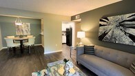 This single-floor residence offers a thoughtfully designed open layout that seamlessly connects the living, dining, and kitchen areas. The living room is a welcoming focal point, featuring rich dark hardwood-style flooring that contrasts beautifully with the warm sage green accent walls. A glass-top coffee table and contemporary sofa create an inviting seating arrangement, while a stylish table lamp adds a warm ambient glow. Wall art provides a decorative touch that enhances the modern aesthetic. A textured ceiling adds architectural character throughout the space. The kitchen is visible from the living area, offering white cabinetry paired with black appliances, including a full-size refrigerator, creating a classic two-tone look. The adjacent dining area is accessible through an open doorway, maintaining a sense of flow and connectivity between the rooms. White baseboards and trim provide clean, crisp definition against the colored walls. The overall ambiance is cozy yet modern, with a neutral and earthy color palette that creates a calm, livable atmosphere suitable for everyday comfort and entertaining alike.