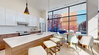 This compact yet thoughtfully designed single-floor space exemplifies contemporary urban design. The layout integrates a fully equipped kitchen with an open dining and lounge area, maximizing functionality within an efficient footprint.

The kitchen is a standout feature, showcasing high-gloss white upper cabinetry paired with warm wood-toned lower cabinets, creating a striking two-tone aesthetic. A large white quartz island serves as the focal point, accompanied by white upholstered stools with gold metallic frames. Stainless steel appliances, including a French door refrigerator and a range, provide a polished, professional finish. Black pendant lights with gold interiors hang above the island, adding a modern decorative touch.

Floor-to-ceiling black-framed windows span a full wall, flooding the space with abundant natural light and offering direct views of the surrounding urban streetscape. The marble-effect tile flooring enhances the overall sense of brightness and sophistication.

The dining area features a statement table with a marble-style top and gold legs, surrounded by contemporary white upholstered chairs. Lush potted greenery adds organic warmth to the otherwise sleek, minimalist interior. The soaring ceiling height amplifies the sense of openness throughout the space.