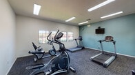 Located at 4970 Camino Delgado in Tucson, Arizona, this single-room fitness center offers a well-organized and functional workout environment. The space is equipped with a variety of cardio machines, including multiple treadmills, an elliptical trainer, and stationary bikes, catering to a range of exercise preferences. Durable, patterned carpet tile flooring covers the entire room, providing both comfort and stability underfoot during workouts.

A full-length mirror spans one wall, visually expanding the room and allowing users to monitor their form while exercising. Opposite the mirror, a teal accent wall adds a pop of color to the otherwise neutral interior, and a wall-mounted flat-screen television provides entertainment during workouts. A row of gridded windows lines another wall, flooding the space with natural light and offering pleasant views of the surrounding desert landscaping, including colorful blooming shrubs and native vegetation.

Overhead, recessed fluorescent lighting ensures the room is consistently well-lit. A sanitizing station is conveniently mounted near the entry door, which connects to an adjacent common area. Free weights, including dumbbells, are also available for strength training. The overall ambiance is clean, practical, and inviting, making it a convenient amenity for residents or guests seeking an accessible on-site fitness facility.