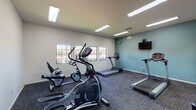 Located at 4970 Camino Delgado in Tucson, Arizona, this single-room fitness center offers a well-organized and functional workout environment. The space is equipped with a variety of cardio machines, including multiple treadmills, an elliptical trainer, and stationary bikes, catering to a range of exercise preferences. Durable, patterned carpet tile flooring covers the entire room, providing both comfort and stability underfoot during workouts.

A full-length mirror spans one wall, visually expanding the room and allowing users to monitor their form while exercising. Opposite the mirror, a teal accent wall adds a pop of color to the otherwise neutral interior, and a wall-mounted flat-screen television provides entertainment during workouts. A row of gridded windows lines another wall, flooding the space with natural light and offering pleasant views of the surrounding desert landscaping, including colorful blooming shrubs and native vegetation.

Overhead, recessed fluorescent lighting ensures the room is consistently well-lit. A sanitizing station is conveniently mounted near the entry door, which connects to an adjacent common area. Free weights, including dumbbells, are also available for strength training. The overall ambiance is clean, practical, and inviting, making it a convenient amenity for residents or guests seeking an accessible on-site fitness facility.