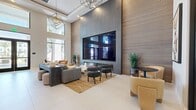 Located in Redlands, CA, this thoughtfully designed single-floor communal space offers a compelling blend of contemporary architecture and upscale finishes across two distinct areas. The main lounge commands immediate attention with its soaring double-height ceilings, large windows that flood the interior with natural light, and views of palm trees beyond. A floor-to-ceiling accent wall clad in textured vertical slat paneling anchors the room and frames a large-format multi-panel display screen. Geometric pendant chandeliers add dramatic lighting, while a long linear fireplace with crystal media creates a warm, inviting focal point that visually separates the lounge into intimate seating zones. Plush sofas, modern accent chairs in warm camel tones, a marble-top coffee table, and a jute area rug contribute to the refined yet approachable atmosphere. The adjoining kitchen is equally impressive, featuring dark charcoal cabinetry with brushed gold hardware, crisp white countertops, a professional-grade stainless steel range, and a bold black-and-white geometric patterned backsplash that extends to the ceiling. A large stainless steel refrigerator and generous prep surfaces complete this highly functional culinary space, which also provides direct access to the outdoors through a glass-panel entry door.