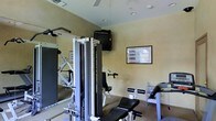 This single-floor fitness space is thoughtfully designed to accommodate a well-rounded exercise regimen. The room features a multi-station cable strength training machine as its centerpiece, paired with an adjustable workout bench ideal for a variety of strength exercises. A wall-mounted flat-screen television provides entertainment during workouts, while a framed exercise chart on the wall offers guidance for training routines. A leg curl/extension bench adds further versatility to the available equipment. A large wall mirror enhances the sense of openness and allows users to monitor their form during exercise. The room is finished with neutral, warm-toned walls and carpeted flooring, contributing to a functional yet comfortable atmosphere. Recessed lighting and a ceiling fan ensure the space remains well-lit and ventilated during use. The overall layout is efficient and practical, making effective use of the available floor space to create a private, fully functional fitness environment suitable for both strength training and general conditioning.