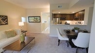 This 3-floor residence offers a thoughtfully designed open-concept main living area that integrates the living room, dining space, and kitchen into a cohesive and functional layout. The living area features plush neutral carpeting and warm, creamy walls that create a welcoming atmosphere, complemented by natural light filtering in through a nearby window. A framed botanical artwork adds a decorative focal point to the space.

The dining area sits adjacent to the living room, accommodating a dark-toned dining table with contemporary white chairs. It flows directly into the kitchen, which is defined by rich dark wood cabinetry, recessed lighting, and stainless steel appliances including a refrigerator and microwave. A peninsula with a light-colored countertop provides both workspace and a natural visual boundary between the kitchen and dining area.

A staircase visible beyond the main living space connects to the upper and lower floors, suggesting generous vertical living across all three levels. The overall ambiance is clean, modern, and inviting, with consistent neutral tones throughout.