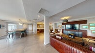 Located at 151 Southeast 3rd Avenue in Delray Beach, Florida, this single-floor space offers a thoughtfully designed, open-concept layout with a cohesive blend of modern and eclectic interior design. Large-format tile flooring flows throughout the main areas, lending a clean, continuous aesthetic.

The kitchen is a standout feature, showcasing warm wood cabinetry with textured panel finishes, sleek white quartz countertops, a center island with under-lit storage, and open shelving flanking a central mirror. Industrial-style pendant lighting adds character overhead.

The living room is anchored by a bold exposed brick accent wall housing a large flat-screen TV, complemented by a statement leather sofa, decorative pendant fixture, and curated artwork. Large sliding glass doors invite natural light and provide views of an outdoor patio area.

Two distinct dining spaces serve different functions — one features a casual bar-height setup, while the other offers a round gaming-style table surrounded by leather chairs beneath an elegant drum chandelier, with windows framing lush outdoor greenery.

A compact secondary area with a vivid red painted brick wall and industrial Edison bulb lighting adds a creative, artistic touch to the space. Overall, the layout balances functionality with bold, expressive design throughout.