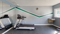 This compact yet fully outfitted fitness center is designed to support a wide range of workout routines within a single, efficiently organized space. The room features gray walls accented by bold green diagonal stripes, a textured ceiling, and durable dark rubber flooring throughout — a practical and visually cohesive design well-suited for an active environment.

The cardio area includes a Spirit treadmill and an elliptical trainer, positioned to take advantage of natural light streaming through a row of white-framed windows along one wall. These windows also offer views of an outdoor pool area, adding a refreshing sense of openness to the space. A multi-station cable machine and a recumbent bike round out the strength and low-impact conditioning options.

A dedicated functional training zone features a Smart rack system stocked with medicine balls and stability balls in various sizes, supporting strength, balance, and recovery exercises. Full-length mirrors along one wall enhance the sense of space while allowing users to monitor their form.

The fitness center is accessed through glass double doors that open to a landscaped outdoor walkway. A security camera, wall-mounted sanitizer station, and posted safety notices reflect a commitment to cleanliness and user safety. A ceiling fan and HVAC vent contribute to a comfortable training environment year-round.