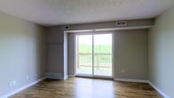 This single-floor unit offers a well-organized layout that makes efficient use of its space. The living area is the centerpiece of the unit, featuring warm hardwood flooring that extends throughout the main living spaces, neutral-toned walls, and a textured ceiling. A large sliding glass door floods the room with natural light and provides direct access to a private balcony, which overlooks a lush, tree-lined landscape — a standout feature that brings a sense of openness and connection to the outdoors.

The kitchen is situated adjacent to the living area and is equipped with a range and essential cabinetry, offering a practical and accessible cooking space. The transition between the kitchen and living room maintains an open, airy feel throughout the common areas.

The bathroom is tucked to one side of the unit and includes a toilet, sink, and tub or shower, all within a clean and compact layout. Additional utility is provided by what appears to be a dedicated entry or storage area near the front of the unit.

Overall, the space presents a straightforward and functional design, ideal for those seeking a manageable, low-maintenance living environment with the added benefit of natural light and pleasant outdoor views accessible from the main living area.