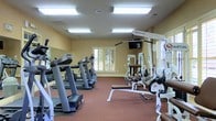 This fitness room offers a practical and well-organized workout environment designed to support a range of exercise needs. The space is finished with warm yellow walls and durable red flooring, creating an energetic yet comfortable atmosphere. Large wall-mounted mirrors along one side of the room visually expand the space and allow users to monitor their form during workouts. Natural light filters in through a shuttered window, complementing the overhead fluorescent lighting to keep the room bright and well-lit throughout the day. The cardio section is equipped with multiple treadmills and an elliptical machine, providing ample options for aerobic exercise. On the strength training side, a Quantum multi-station cable machine and a weight bench are positioned to offer a variety of resistance exercises. A wall-mounted flat-screen television adds convenience for entertainment during workouts. The room is thoughtfully arranged to allow comfortable movement between equipment without overcrowding, making it a functional and inviting space for daily fitness routines.
