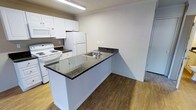 This single-floor space is designed with practicality and clean aesthetics in mind. The kitchen is a standout feature, showcasing white cabinetry that extends to the upper walls, providing ample storage and a bright, cohesive look. Dark granite countertops offer a striking contrast against the white cabinets and appliances, adding a touch of elegance to the functional space. A peninsula island serves as the centerpiece of the kitchen, featuring the same granite surface and a stainless steel sink, making it ideal for meal preparation and casual use. White appliances, including a refrigerator, electric range, and over-the-range microwave, are seamlessly integrated into the cabinetry layout. The flooring transitions between warm hardwood-style planks and a lighter tone in adjacent areas, lending visual interest and definition to different zones within the space. Overhead fluorescent lighting ensures the kitchen remains well-lit throughout the day. The neutral wall tones throughout create a clean, open atmosphere that complements the overall design. The layout is efficient and thoughtfully arranged, making excellent use of the available space on a single level.