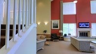 This single-floor space makes a strong first impression with its dramatic double-height ceiling, which lends an open, airy quality to the main living area. The interior design balances warm and neutral tones, pairing rich terracotta-red accent walls with softer cream and beige surfaces to create a visually dynamic yet inviting ambiance. A white-railed staircase is positioned prominently, serving as an architectural focal point while adding vertical interest to the space. The main seating area is thoughtfully arranged with upholstered furniture, including a cushioned sofa and a dark armchair, offering comfortable conversation areas. Wall-mounted sconces provide warm accent lighting, complementing the abundant natural light that streams in through large windows fitted with roller shades. The tall windows offer views of lush greenery outdoors, further enhancing the sense of connection to the exterior environment. A framed artwork piece and decorative potted plants add curated touches throughout. A curved architectural element near the window area adds a refined design detail. The carpeted flooring throughout contributes to the overall warmth and comfort of the space.