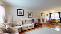 This single-floor home offers a cohesive and comfortable layout connecting several functional living spaces, all unified by warm hardwood flooring. The living room serves as the heart of the home, featuring a plush sectional sofa, accent chairs, and framed artwork adorning the crisp white walls. Natural light filters in through windows dressed with sheer curtains, creating a bright and welcoming atmosphere. A front entryway with a windowed door opens directly into the living space, reinforcing the home's airy feel.

Adjacent to the living room, a dedicated dining area provides a cozy space for meals, anchored by a glass-top round table with rattan chairs and framed by a window with navy drapery offering pleasant outdoor views. The kitchen is equipped with warm oak cabinetry, a gas range, dishwasher, and ample counter space, complemented by a small eat-in table for casual dining.

The two bedrooms are comfortable and well-lit, each featuring hardwood floors, windows with natural views, and sufficient space for bedroom furnishings. The bathroom includes tile flooring, a tub/shower combination, a vanity with storage, and a window that allows natural light to brighten the space. Overall, this home presents a practical and inviting single-level layout with consistent, warm finishes throughout.