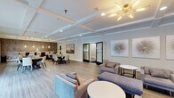 Located at 101 Steuben Dr in Albany, NY, this Heritage Village amenity space offers a generously sized, single-floor layout designed for community gathering and recreation. The open-concept living and dining area serves as the heart of the space, featuring a coffered ceiling with a striking sputnik chandelier, warm wood-look flooring, and an abundance of natural light streaming through large windows dressed with linen drapes and roller shades. Comfortable seating arrangements with upholstered chairs and accent tables create inviting lounging zones, while a dedicated dining area accommodates multiple seating groups beside a textured tile accent wall. Curated wall art, including a trio of starburst canvases, adds a polished, contemporary aesthetic throughout. A welcoming entry foyer greets visitors with bold navy accent chairs, a glass coffee table, and a stylish textured wall panel. The kitchen is sleek and functional, outfitted with navy cabinetry, marble-look countertops, stainless steel appliances, and pendant lighting. The fitness center is well-appointed with treadmills, strength equipment, floor-to-ceiling mirrors, and soaring ceilings with natural light, all beneath elegant cage-style pendant fixtures. Multiple hallways connect each zone seamlessly throughout this well-appointed community facility.