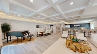 This sophisticated two-story home showcases a seamless blend of classic architectural elements and contemporary design. The expansive main living area immediately commands attention with its stunning coffered ceiling, recessed lighting, and ambient cove illumination, creating a bright and airy atmosphere. Light hardwood flooring flows throughout, complementing the neutral palette of the space. A grand piano anchors one corner, while a formal dining area and comfortable seating zones define the open-plan layout. Adjacent to the living area, a wet bar or kitchenette features striking marble-veined stone countertops and backsplash with dark wood cabinetry, adding a refined focal point. A dramatic ironwork staircase with an ornate crystal chandelier serves as an architectural centerpiece connecting the two floors. The upper level houses well-appointed bedrooms bathed in natural light, each featuring tray ceilings with subtle lighting details and light hardwood floors. The bathrooms are finished to a high standard, with large-format stone tiles, glass-enclosed walk-in showers with rain heads, and backlit vanity mirrors. A wide, well-lit corridor with clean white millwork connects the private rooms. Throughout the home, the consistent use of neutral tones, quality materials, and thoughtful architectural detailing creates a cohesive and refined living environment.