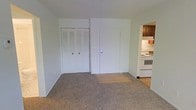 This single-floor space offers a straightforward and functional layout. The main living area is finished with neutral-toned carpet, providing a comfortable underfoot feel throughout the room. White walls keep the space bright and versatile, lending itself well to a variety of décor styles.

A set of bifold louvered closet doors lines one wall, offering practical built-in storage. Adjacent to the closet is a solid entry door with a standard lock, featuring a small transitional area of hard flooring at the threshold where the carpet meets a smooth surface.

Directly accessible from the main living area is a compact kitchen, partially visible through an open passageway. The kitchen includes white appliances — notably a range and oven — along with dark-toned wooden cabinetry above. An under-cabinet range hood is also present, adding functional ventilation to the cooking area.

Overall, the space presents a clean, no-frills environment suitable for everyday living. The open sightline between the living area and kitchen enhances the sense of connectivity, making the layout feel cohesive despite its modest scale.