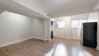This single-floor unit offers a clean, understated design defined by neutral tones, warm hardwood-style flooring, and crisp white trim throughout. The open living area is generously proportioned, benefiting from natural light that streams in through multiple windows fitted with horizontal blinds. Structural columns and subtle level changes add architectural character to the otherwise minimalist layout, while smooth white ceilings and freshly painted walls create a bright, airy atmosphere.

The kitchen is efficiently designed, featuring dark espresso-toned cabinetry — both upper and lower — paired with a light countertop, a stainless steel sink, a gas range, and a dishwasher. A window above the counter allows natural light to filter into the space. The kitchen flows naturally into the main living area, reinforcing the open-concept feel of the unit.

The bathroom is finished in a classic style, with a full soaking tub and shower combination, chrome fixtures, a white vanity, and a distinctive black-and-white octagon mosaic tile floor that adds timeless visual interest. A small window provides ventilation and natural light. The bedroom area is spacious and open, with clean walls and ample room for furnishings. Overall, this space presents a functional and well-maintained interior suited for comfortable everyday living.