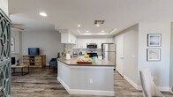 Located in Phoenix, AZ, this 2-bedroom, 1-bathroom home offers a well-appointed single-level layout with modern finishes throughout. The open-concept kitchen and living area serves as the heart of the home, featuring white shaker cabinetry, subway tile backsplash, stainless steel appliances including a gas range, microwave, and refrigerator, and a wraparound peninsula with a built-in sink and neutral countertops. A tray ceiling with recessed lighting adds architectural interest to the kitchen. Wood-style flooring flows seamlessly through the main living areas, complementing the bright, neutral color palette. The living room is anchored by a tiled fireplace and sliding glass doors that invite abundant natural light while providing access to an outdoor space. A ceiling fan enhances comfort throughout the room. Both bedrooms are carpeted and fitted with ceiling fans, offering a quiet retreat. The primary bedroom is generously sized and connects to a spacious walk-in closet with multi-tiered shelving and hanging rods. The full bathroom features a wide vanity with white cabinetry, a large mirror, and wood-style flooring. The overall aesthetic is clean, contemporary, and move-in ready.