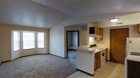 This single-floor residence offers a straightforward, functional layout designed to maximize everyday livability. The open-concept living area is finished with wall-to-wall carpeting and neutral-toned walls, creating a calm and understated atmosphere. A large multi-pane window floods the living space with natural light, enhancing the sense of openness throughout.

The kitchen is positioned adjacent to the living area and features a peninsula countertop that serves as both a workspace and a casual divider between the two zones. Warm wood-toned cabinetry lines the upper and lower sections of the kitchen, complemented by white appliances including an electric range and dishwasher. The kitchen floor transitions to light-toned tile, providing a practical and easy-to-maintain surface.

A doorway visible from the main living area leads further into the residence, suggesting additional rooms beyond the open living and kitchen zone. The textured ceiling and clean wall finishes carry throughout, maintaining a consistent and cohesive interior aesthetic. The overall ambiance is practical and welcoming, with a neutral palette that offers a versatile backdrop for a variety of furnishing styles.